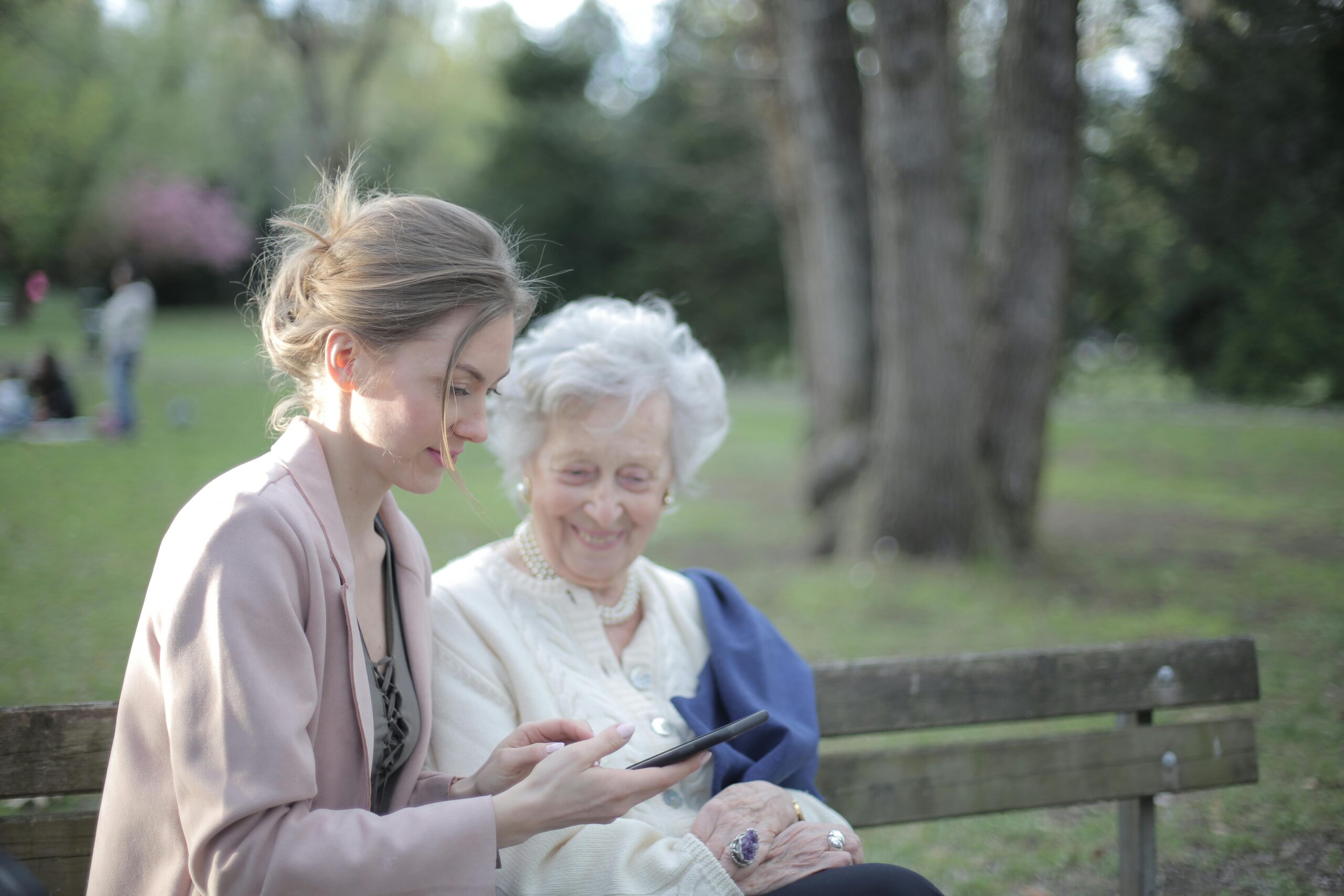 Home Side view of smiling adult female helping aged mom in using of mobile phone while sitting together in park