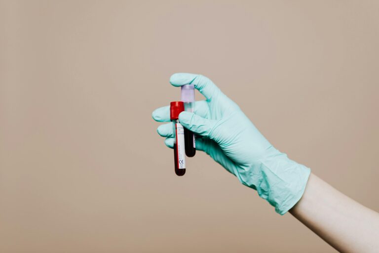 Home Close-up of a gloved hand holding blood test tubes against neutral background.